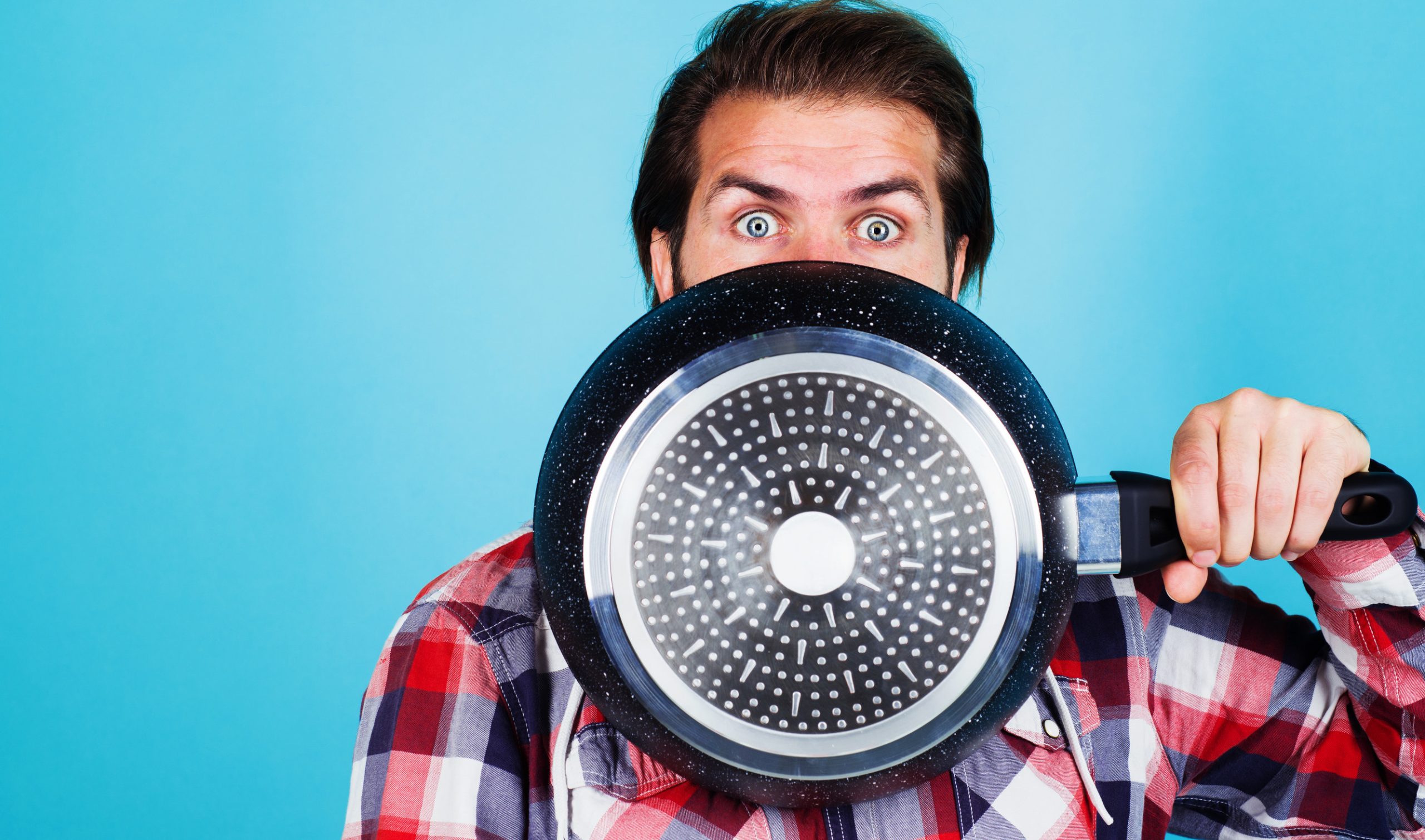 Man holding a large frying pan in front of his face, showcasing kitchen tools for culinary creativity, against a bright blue background.